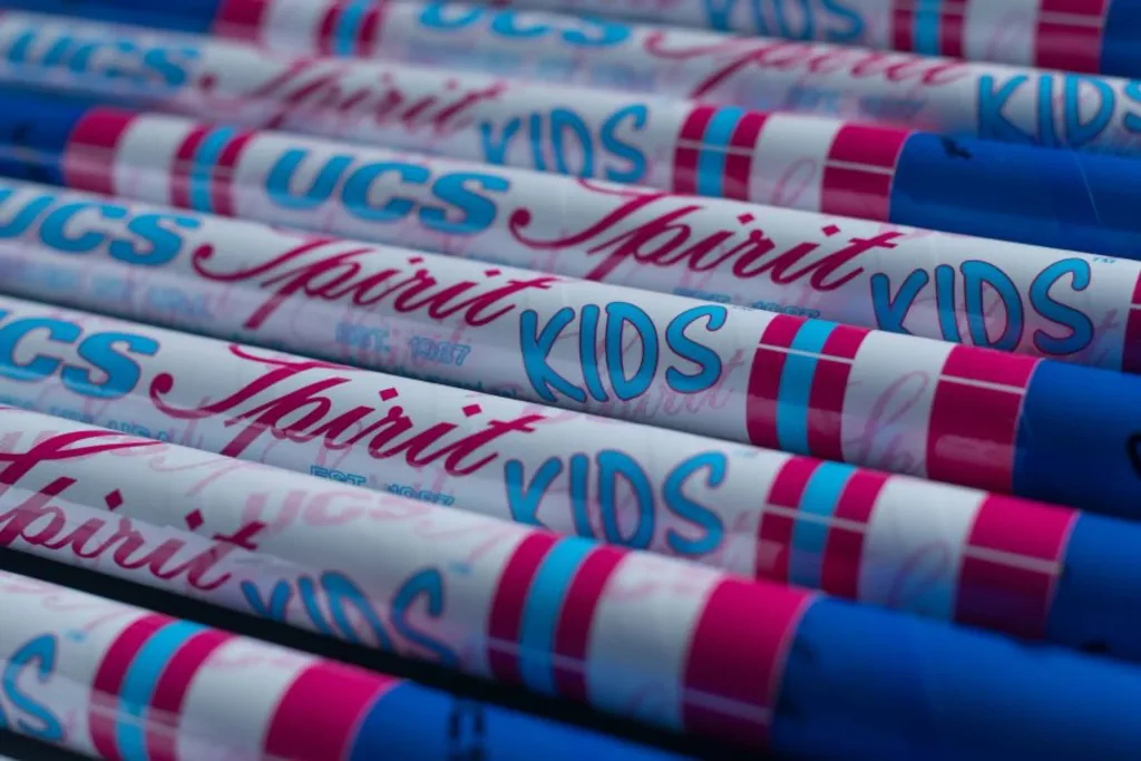 Rows of white junior vaulting poles printed with "UCS", "Spirit", and "KIDS" in blue and pink, with blue end caps.