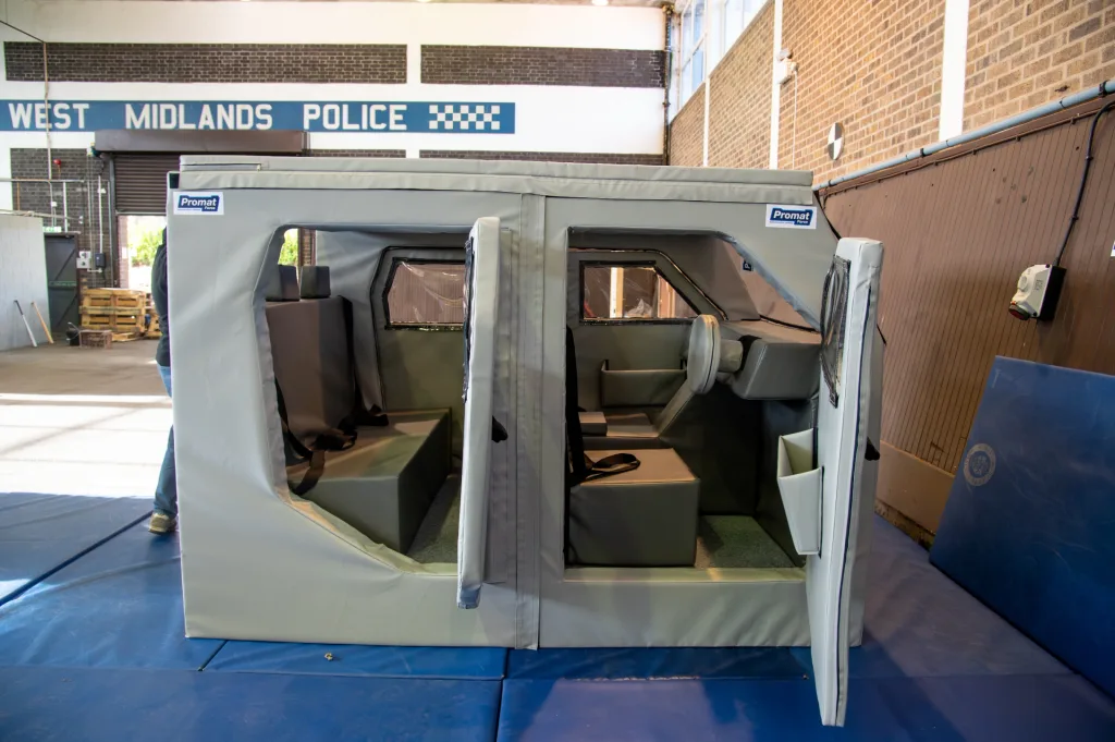 Gray shock-absorbing foam training car with heavy-duty PVC cover, open door revealing seats and steering wheel in a hall with West Midlands Police sign.