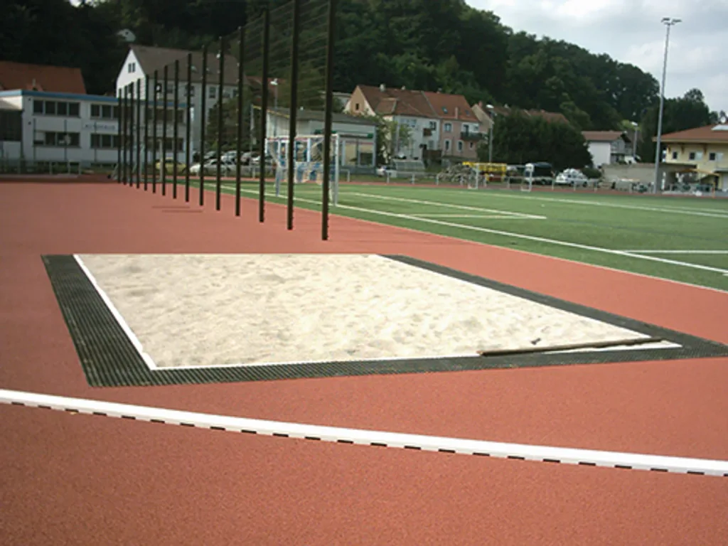 Rectangular long jump sandpit surrounded by black mesh-grating sand traps with a rubber mat and polypropylene base on a red running track.