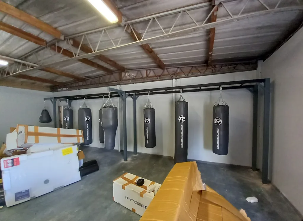 Steel equipment rig with six hanging heavy bags in a warehouse-style gym area and cardboard boxes in the foreground.