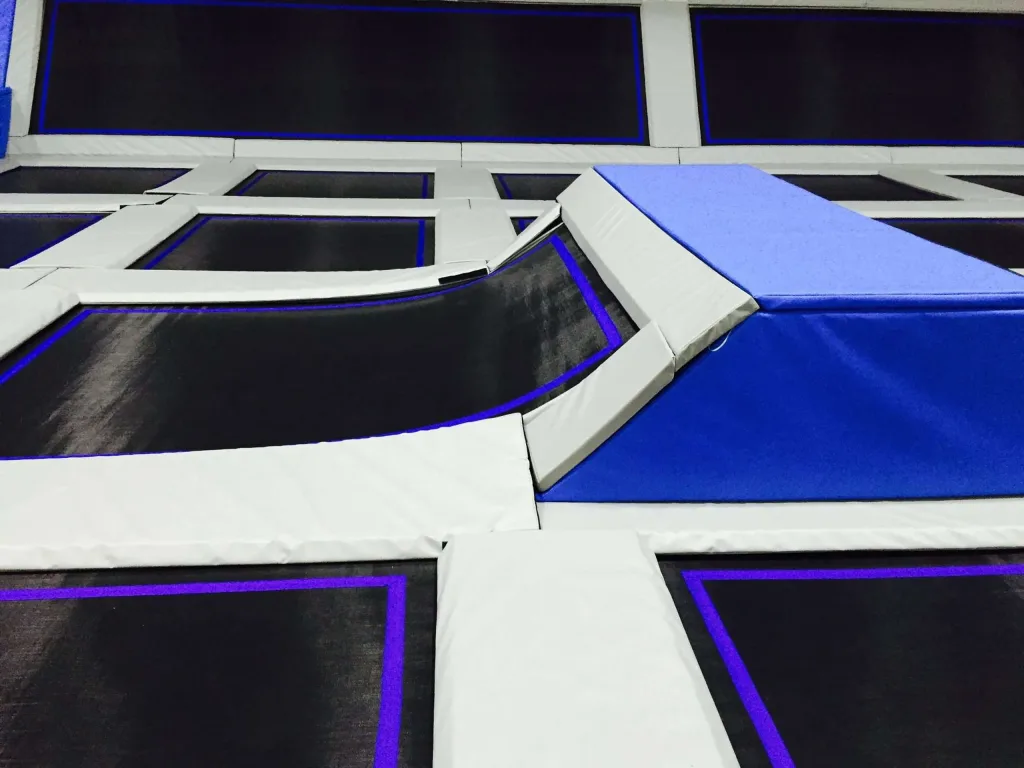 Interconnected trampolines with white PVC frame padding and a blue foam angled landing pad over black jump surfaces with purple trim.