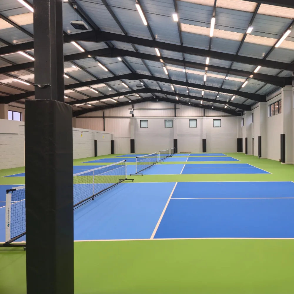 Black padded post in the foreground beside several blue pickleball courts with green surrounds and nets in an indoor metal-roofed hall.