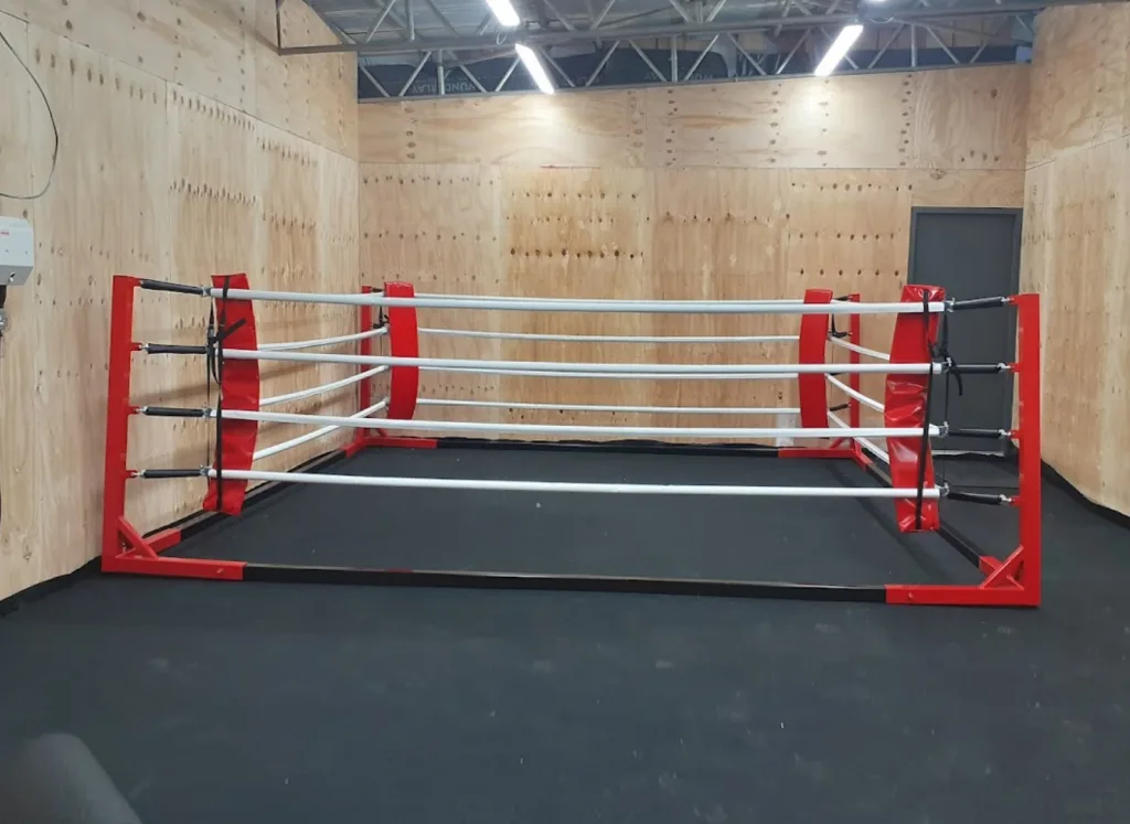 Floor standing ring with powder-coated steel frame, four ropes, corner pads and black canvas inside a plywood-walled training room.