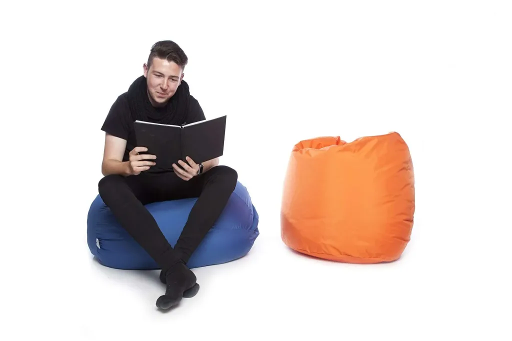Person sitting on a royal blue polyester bean bag reading a book, with an orange polyester bean bag beside on a white background.