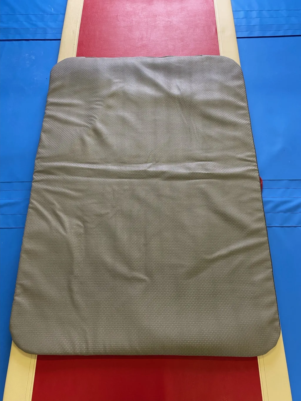 Gray jersey fabric balance beam mat with PU foam core resting on a red and yellow beam atop a blue landing mat.