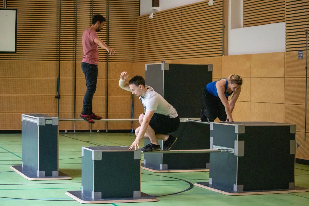 Three people practicing parkour on multiplex birch boxes with high-grip surfaces and metal poles in a gymnasium.