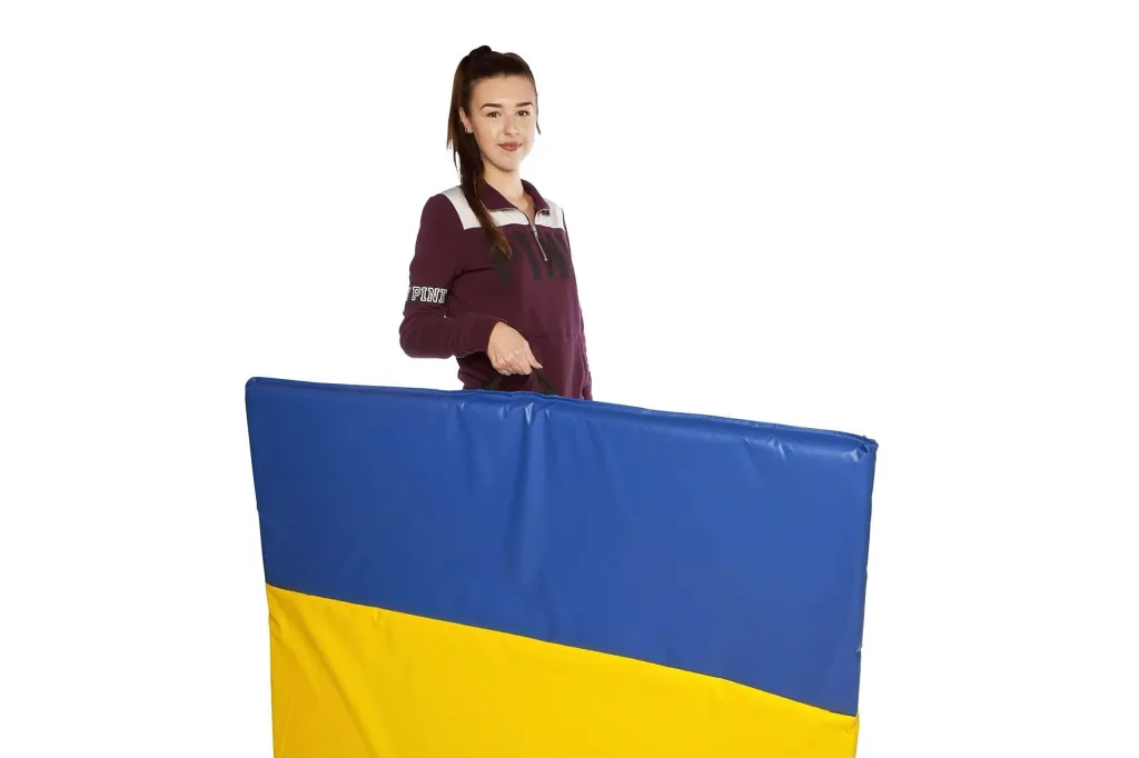 Woman holding a folded giant tumble mat with blue top and yellow bottom panels and a black carry handle, 'PINK' visible on her sleeve.