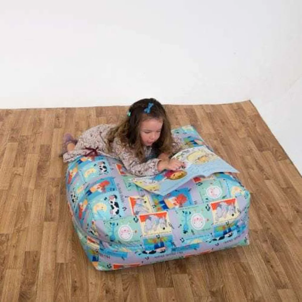 Young girl lying on a square child bean bag with a patterned cotton cover, reading a picture book.