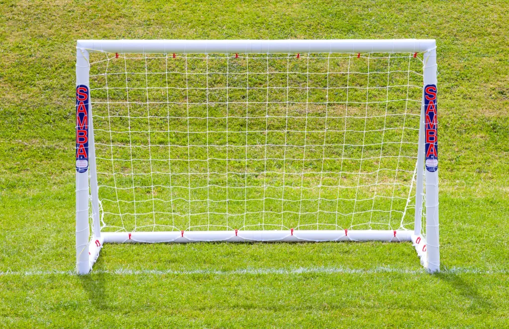 White soccer goal with white net on green grass, vertical label "SAMBA" on both posts.