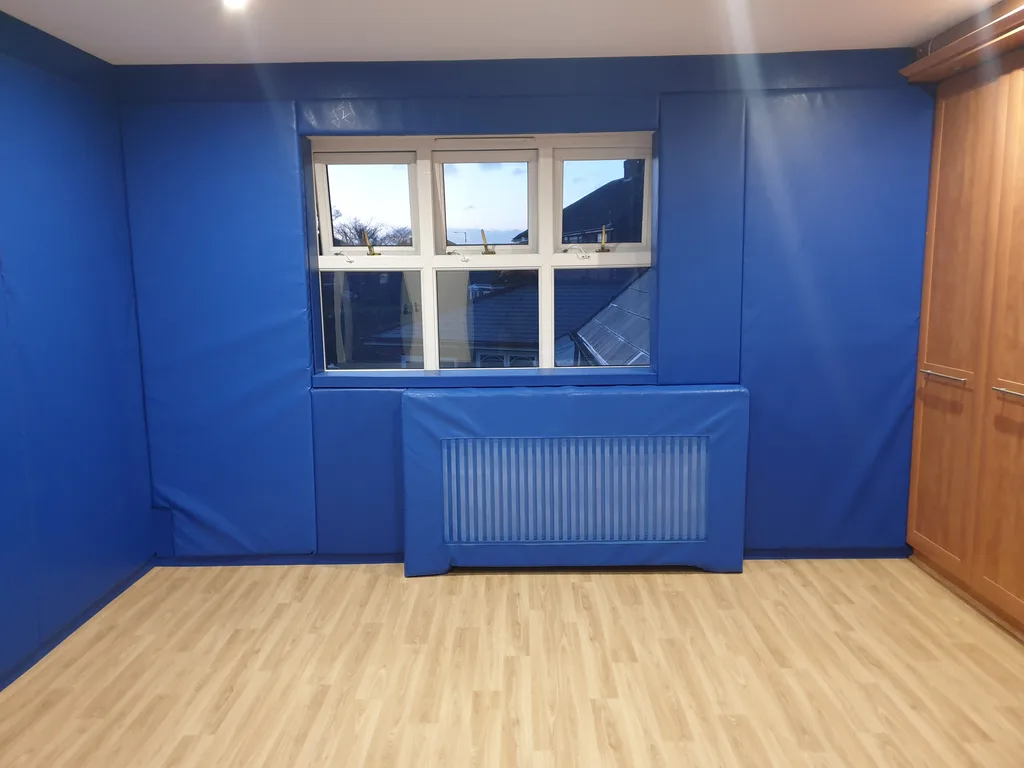 Blue padded radiator cover with a mesh ventilation panel beneath a window, set against blue wall padding in a wood‑floor room.