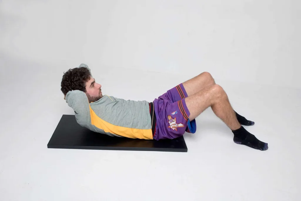 Man lying on a black mat doing a crunch with hands behind his head, wearing a gray and yellow top, purple shorts, and black socks.