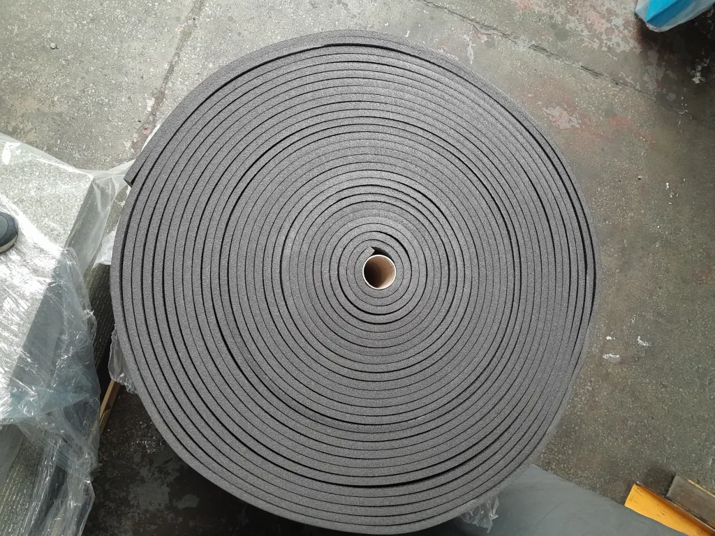 Large dark gray foam roll with concentric spiral layers and a cardboard core resting on a concrete floor beside plastic wrapping.