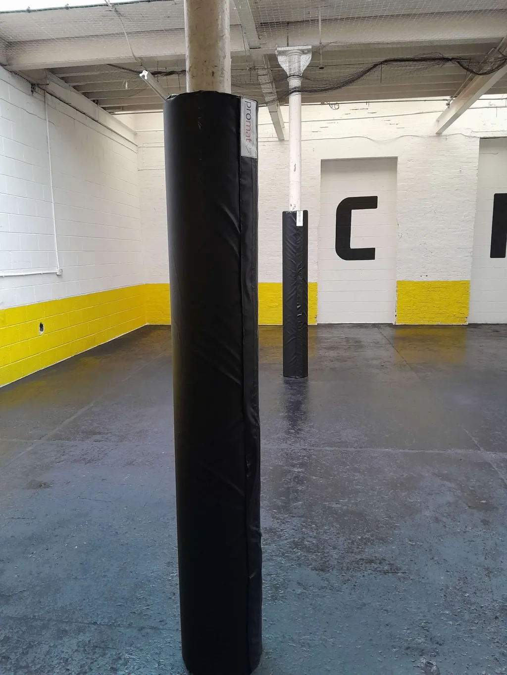 Black round post protector on a white column in a warehouse room with white walls and a yellow lower stripe and a second wrapped post behind.