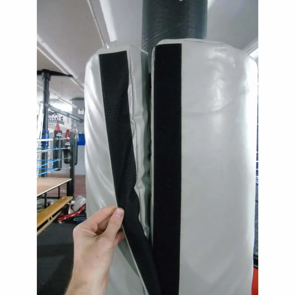 Hand peeling back a black Velcro strip on a light gray round post protector wrapped around a metal post in a boxing gym.