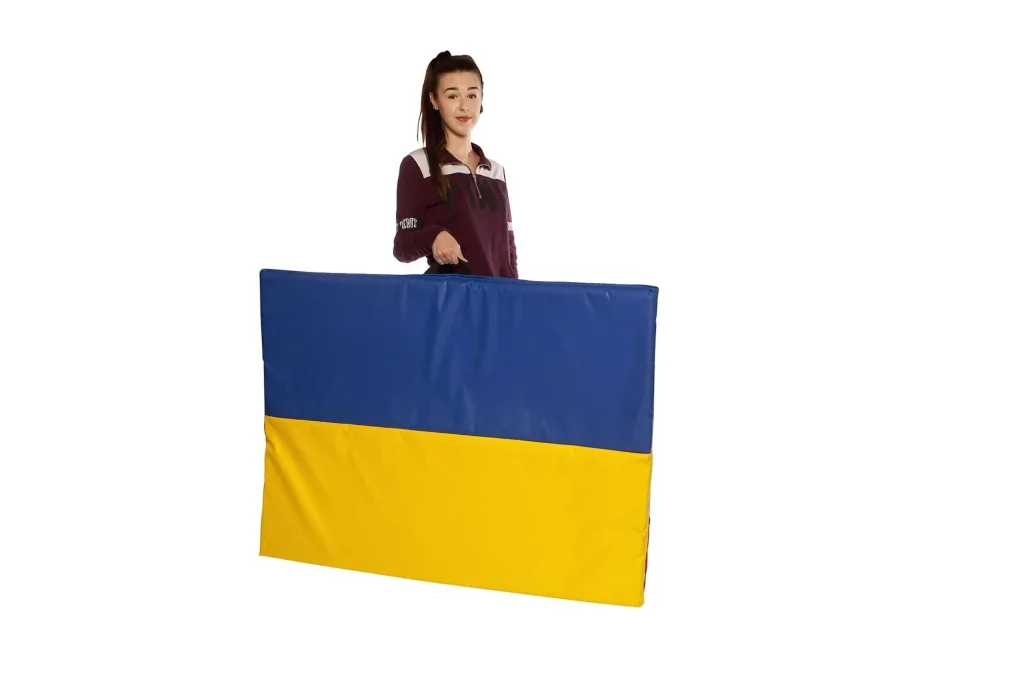 Woman holding a folded giant tumble mat with a blue top and yellow bottom by its carry handle.