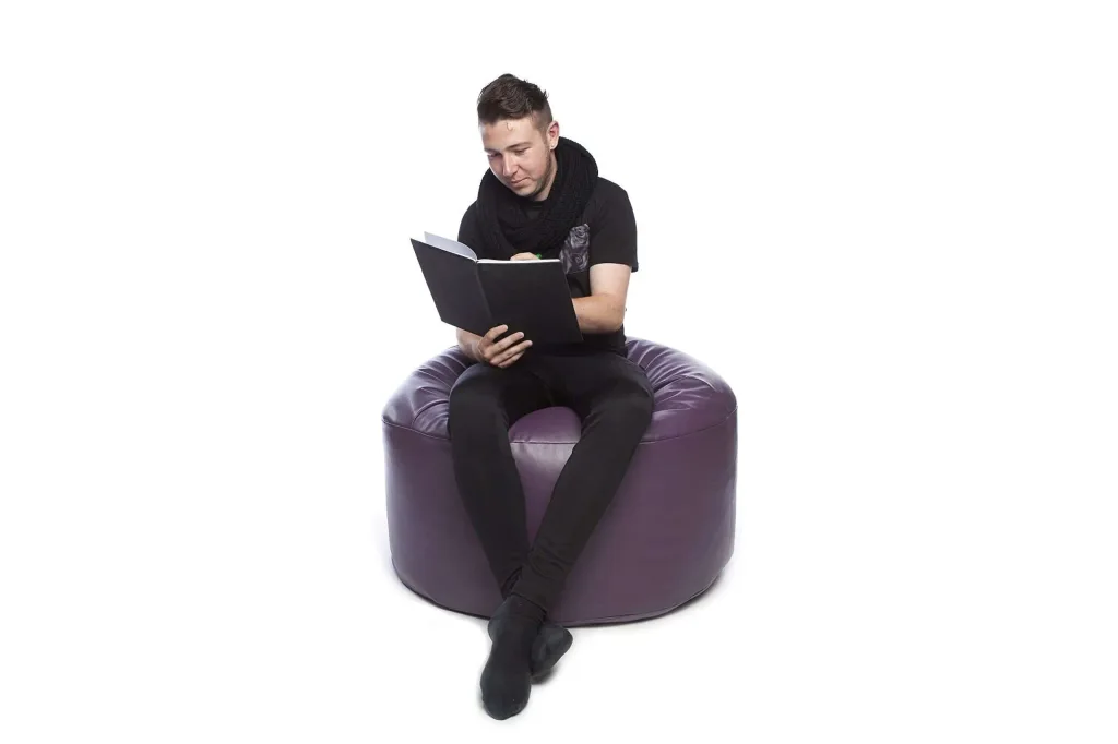Man in black clothing sitting with legs crossed on a purple faux leather round bean bag reading a book.