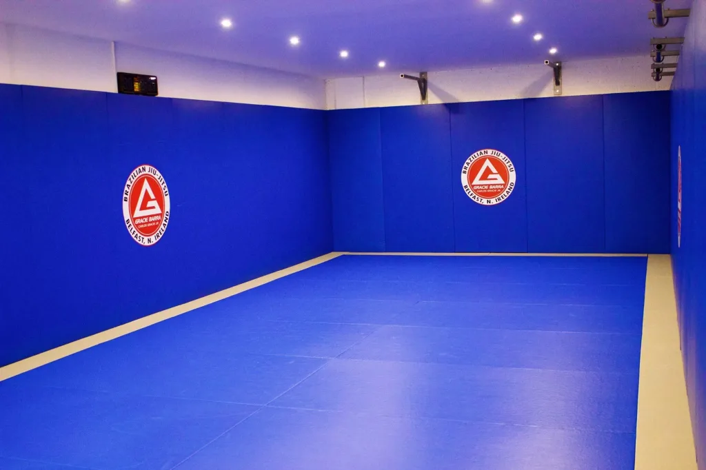 Royal blue polyester PVC jiu-jitsu mat with latex anti-slip base, padded blue wall panels and small circular red-and-white logos.