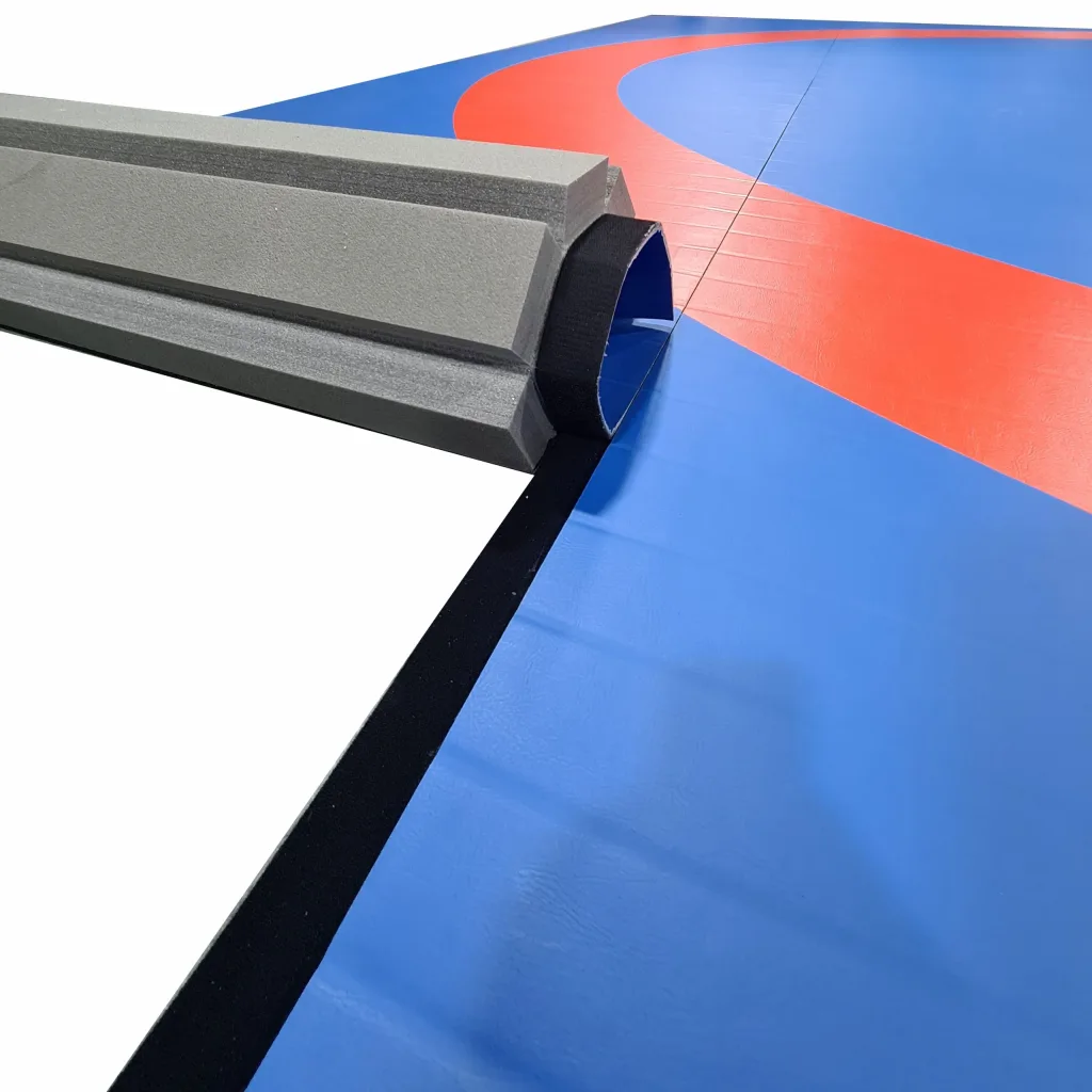 Wrestling roll-out mat with gray foam edge rolled back to reveal a blue PVC surface with a red circle and black seam tape.