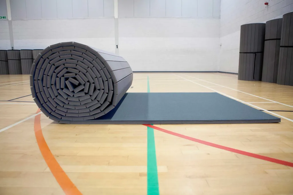 Rolled carpet roll-out mat partially unrolled on a gym floor, showing layered XPE foam core and stacked rolled mats in the background.