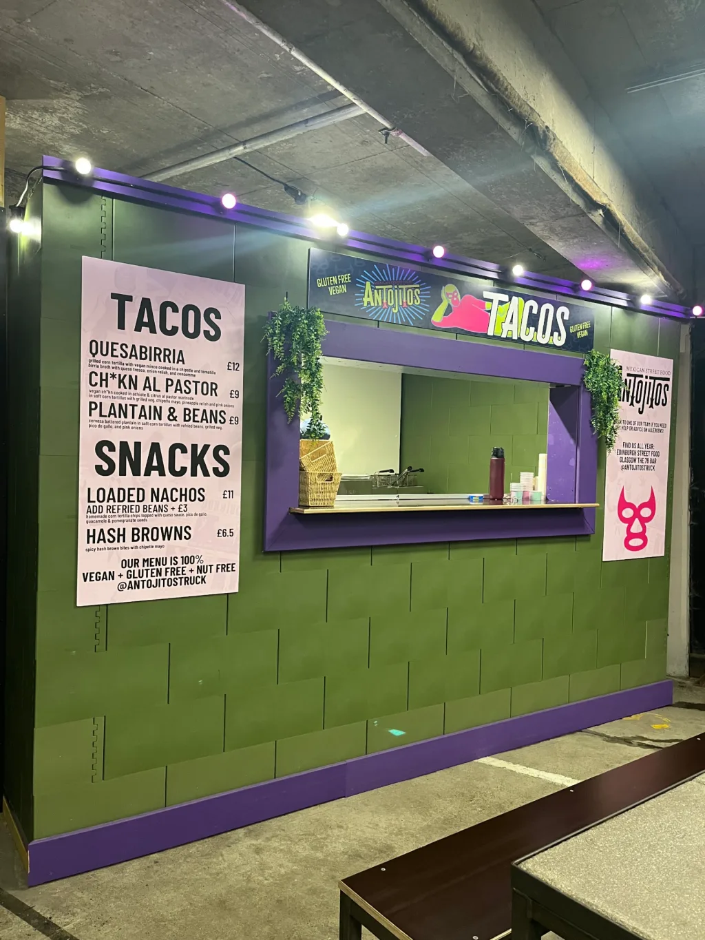 Green modular wall with purple serving window under a concrete ceiling, menu board and large sign reading TACOS.