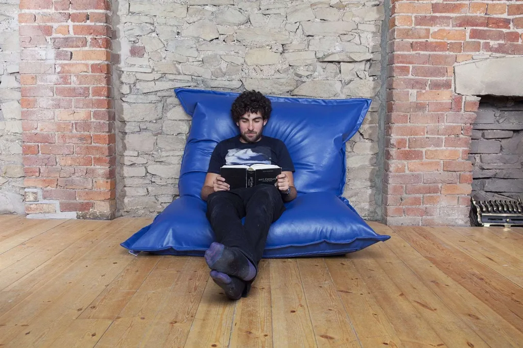 Man sitting cross-legged reading on a royal blue faux leather teen/adult floor cushion against an exposed brick wall on a wooden floor.