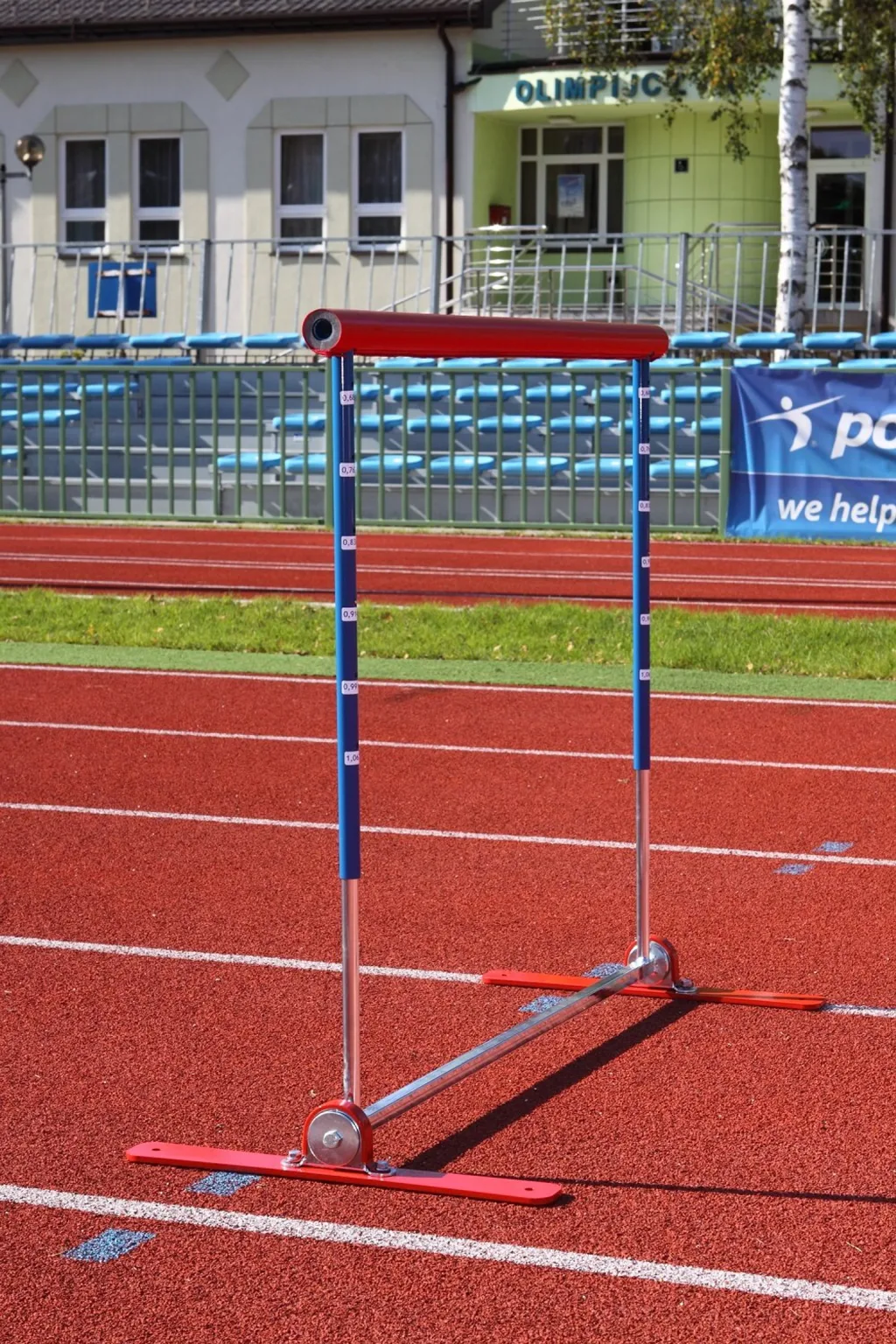 Red-padded hurdle with blue uprights and silver legs on a red track, signage 'OLIMPIJC' and banner 'we help'.