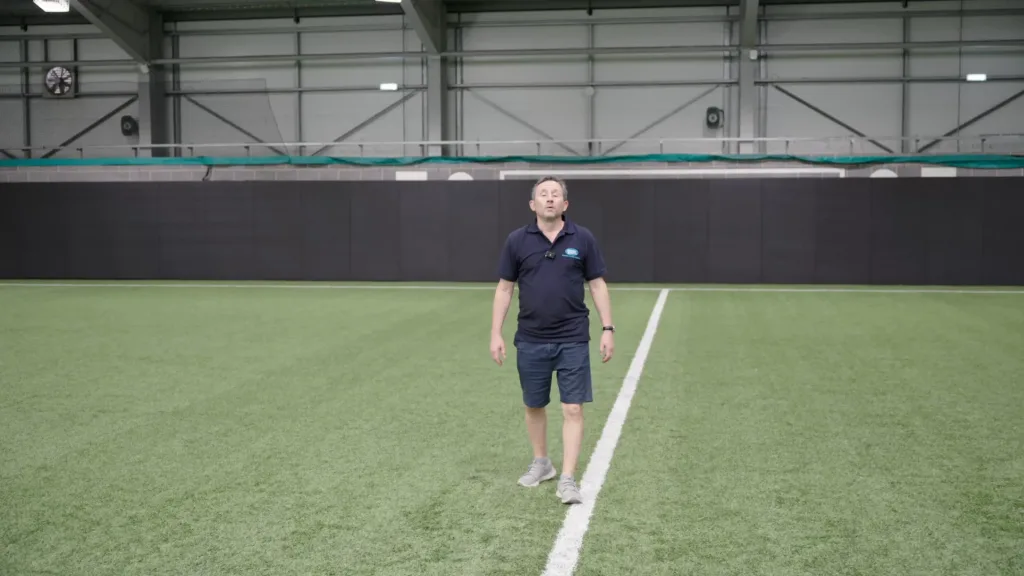 Man in a navy polo and shorts walking on indoor green turf along a white line with a dark padded wall and industrial ceiling behind him.