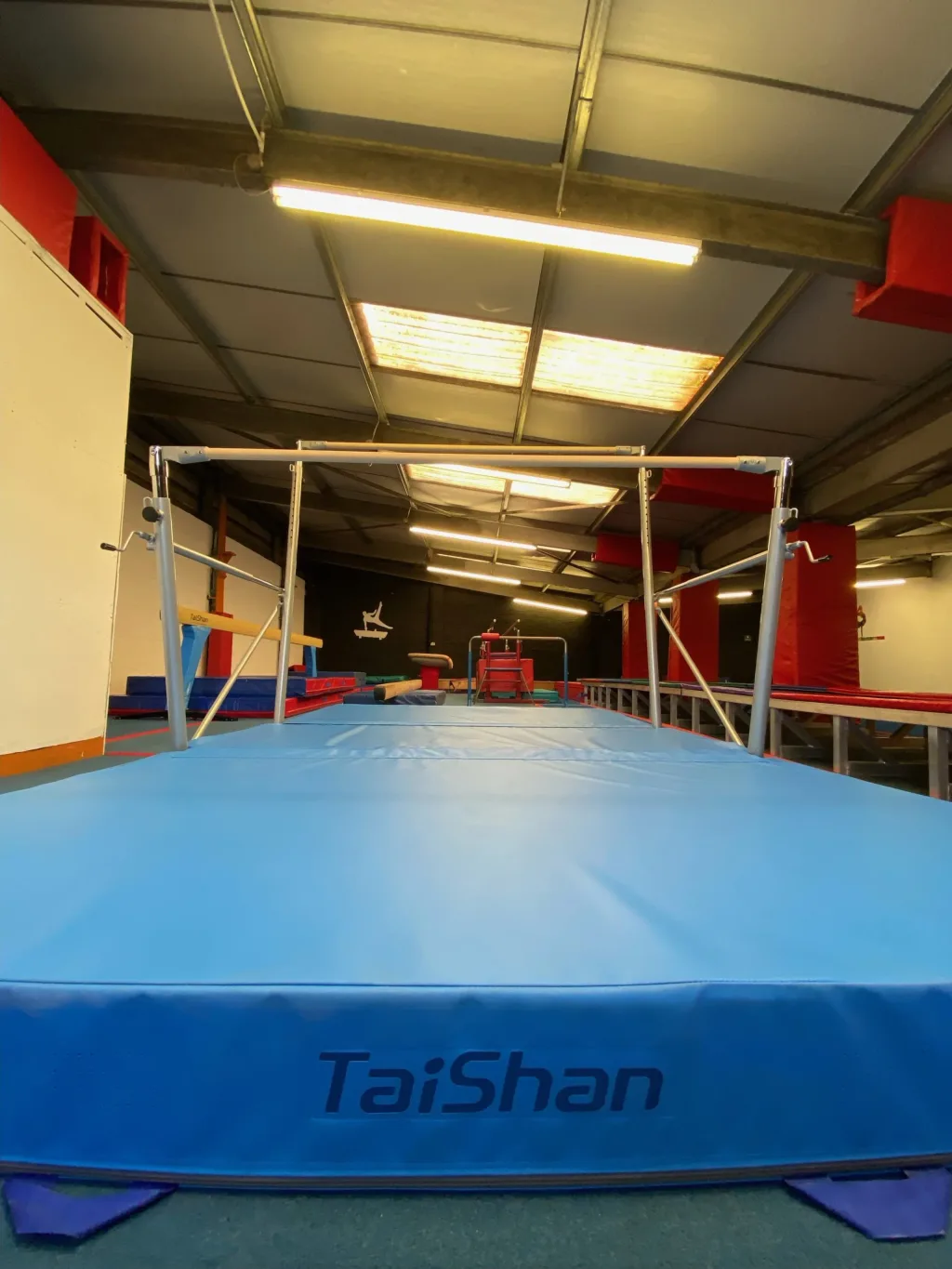 Freestanding uneven bars over a large blue foldable landing mat labeled TaiShan in a gymnasium.
