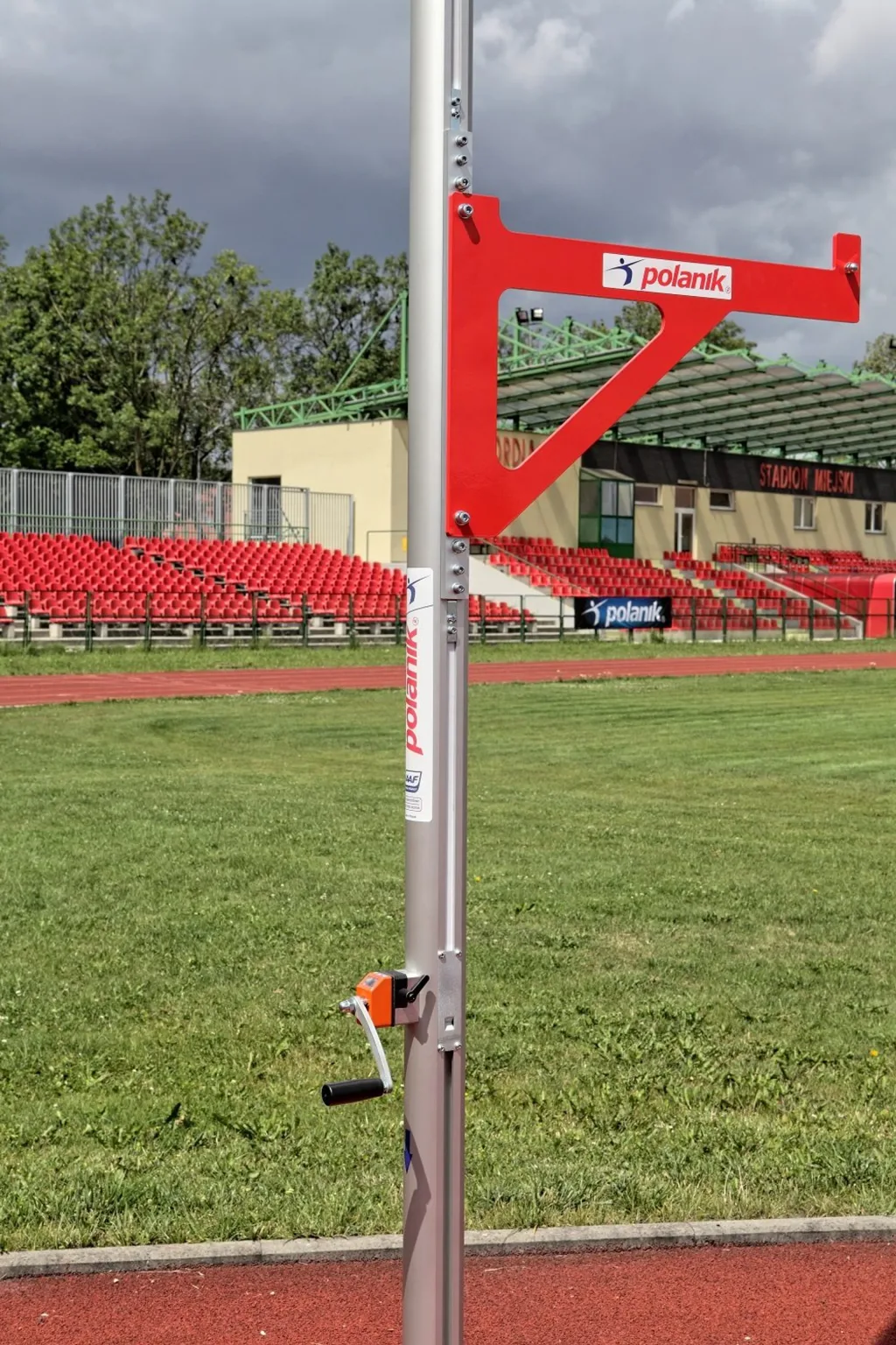 Anodized aluminium pole vault upright with powder-painted aluminium crossbar support on a galvanized-steel wheeled base at a stadium.
