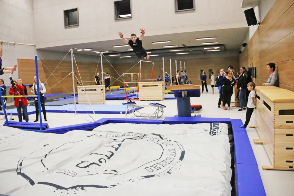 Male gymnast wearing black gloves and shirt leaps airborne over a white crash mat stamped "DENMARK" in a gymnasium with spectators and equipment.