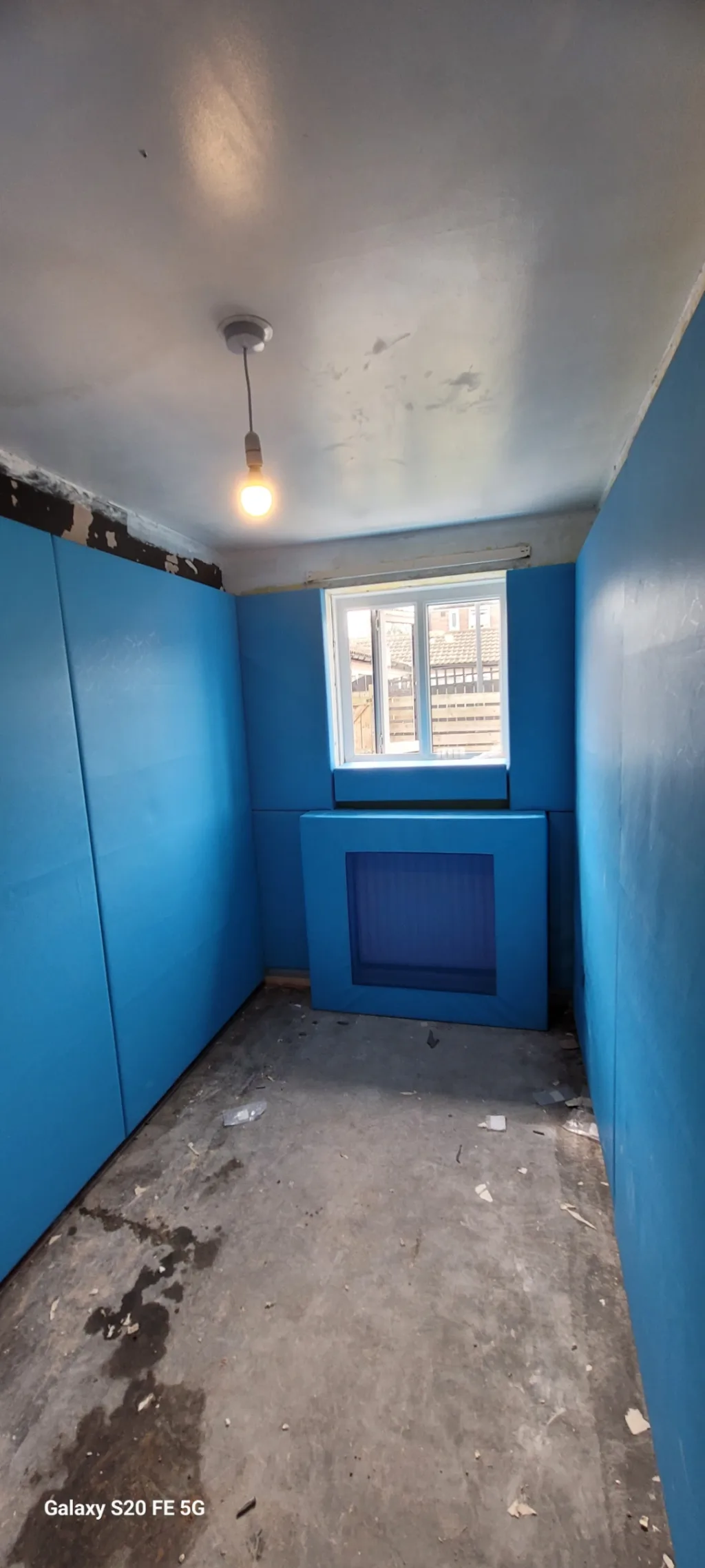 Small padded room with bright blue walls and radiator cover, bare concrete floor, single hanging bulb and window Galaxy S20 FE 5G.