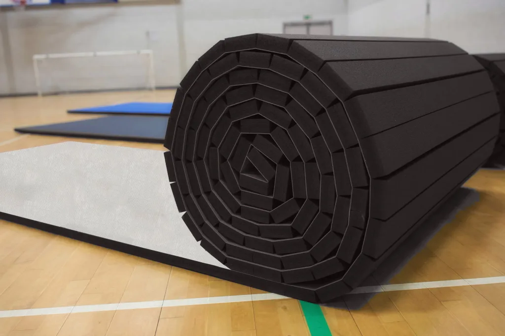 Black hybrid roll-out PVC mat made from high-density cross-link foam, rolled on a gym floor with blue and black mats unrolled in the background.