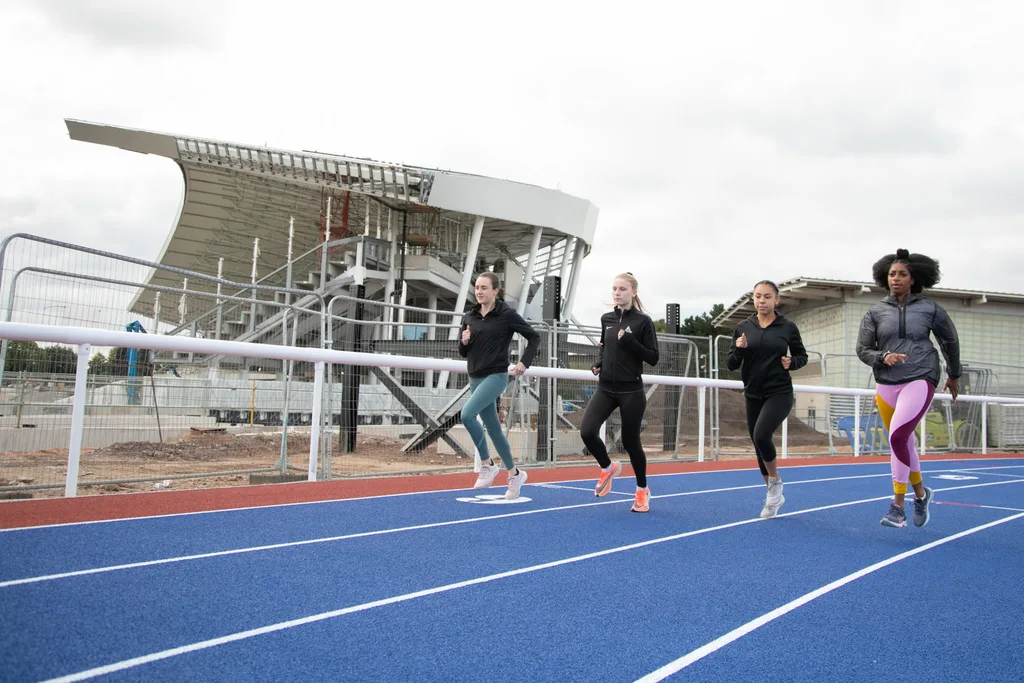 Four women running on a blue track beside a partially built stadium.
