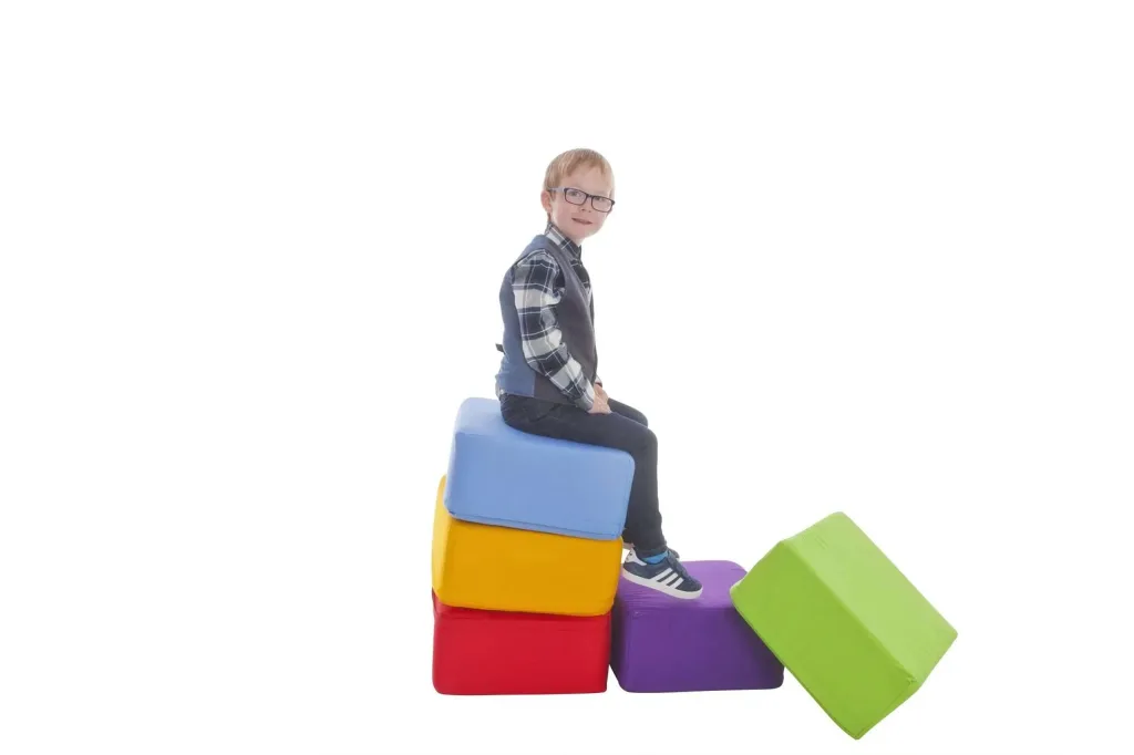 Child with glasses sits on a stack of five cotton-covered square foam seating pods in red, yellow, blue, purple, and green.