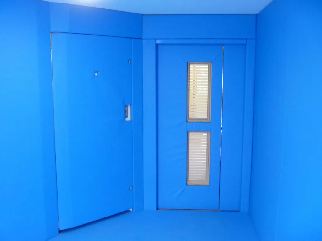 Blue padded room with cushioned walls and floor, a hinged padded door ajar on the left and a padded sliding door with two narrow glazed windows on the right.