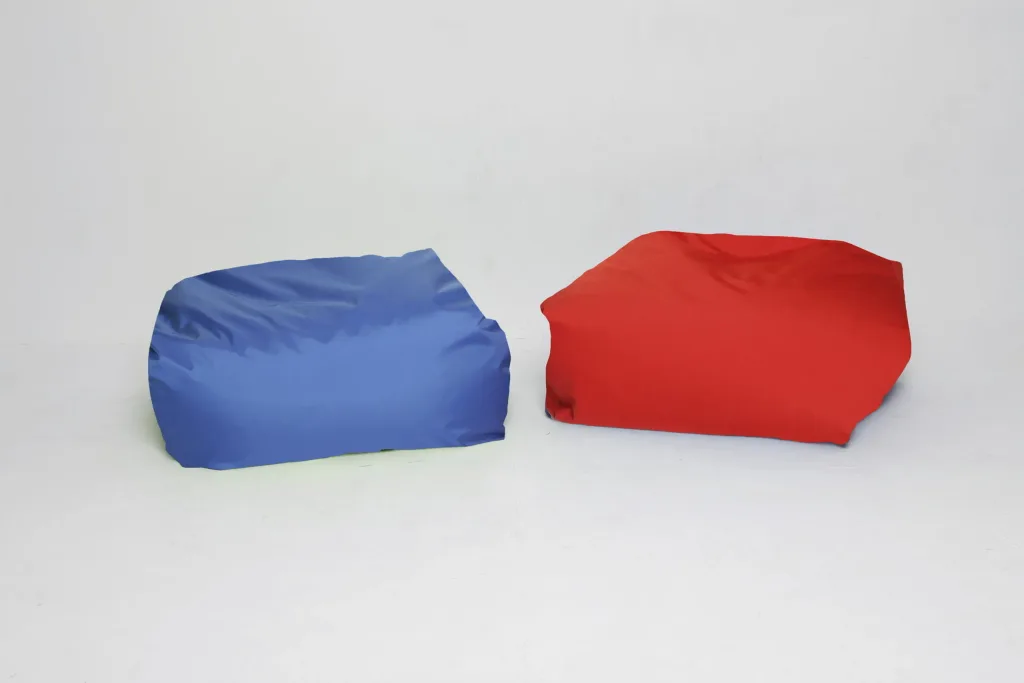 Two child-sized square polyester bean bags, one blue and one red, placed side by side on a white floor against a plain white background.