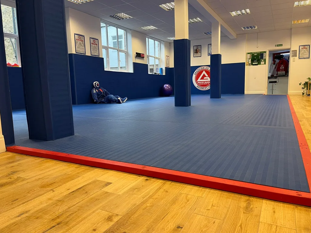 Blue matted training room with red trim, padded columns, a mannequin in a gi by the wall, a purple exercise ball, and a clock reading 9:39.