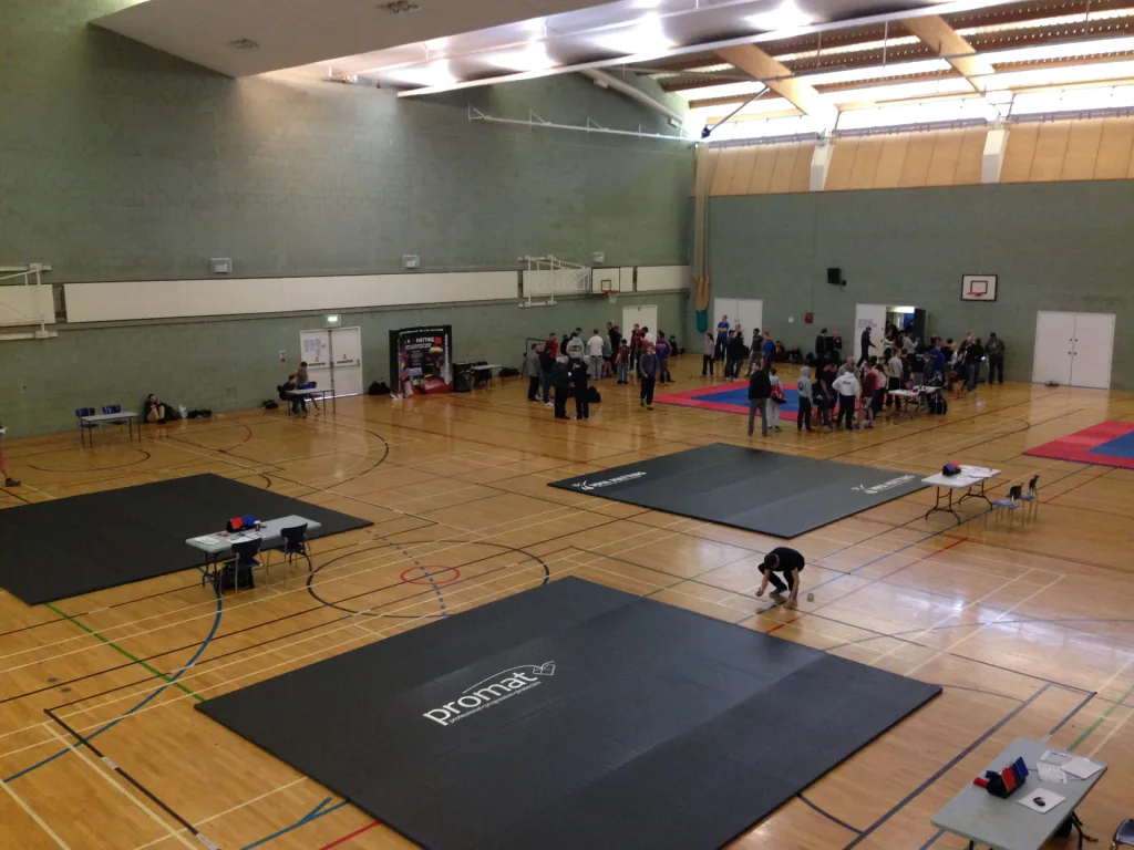 Large dark roll-out martial arts mats labeled promat on a gymnasium floor, with tables and groups of people gathered near other mats.