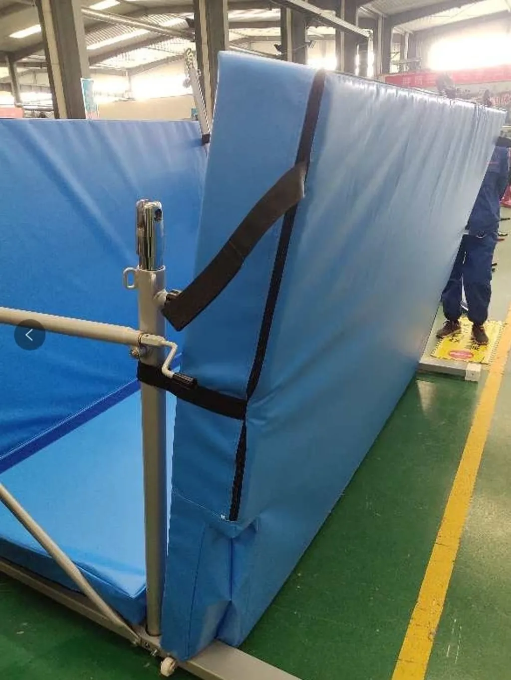 Blue foldable landing mat strapped to a freestanding metal uneven bars frame, showing the adjustment post and horizontal bar.