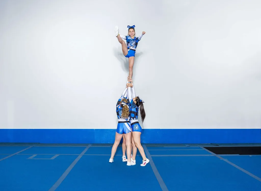 Four cheerleaders on a blue carpet roll out mat perform a stunt, three bases lifting a flyer high with one leg extended.