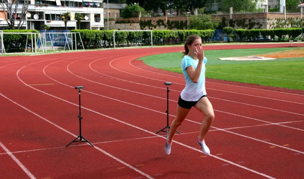 Young woman sprinting mid-stride on a red track passing two tripod-mounted electronic timing gates.