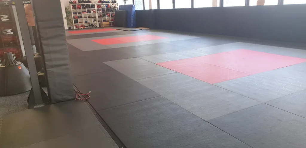 Gym floor covered with black PVC contest mats with red square fighting areas and a padded column at the left.