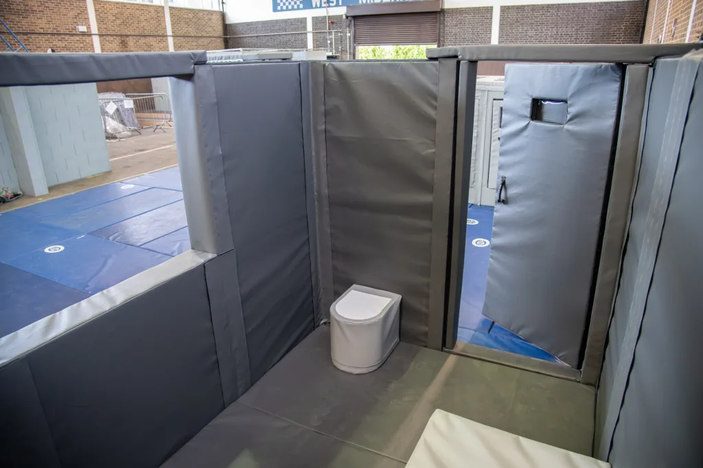 Padded mock custody cell of grey PVC-wrapped foam panels with a foam toilet and an open padded door showing blue training mats outside.