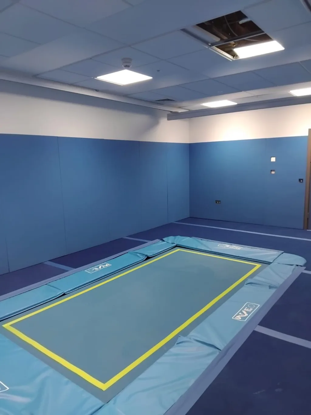 Blue rebound trampoline with a yellow inner border and padded surround in a therapy room with blue wall padding and drop ceiling.