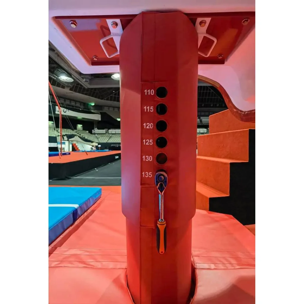 Red vaulting table central column with height holes labeled 110 115 120 125 130 135 and an attached adjustment wrench, arena seating behind.