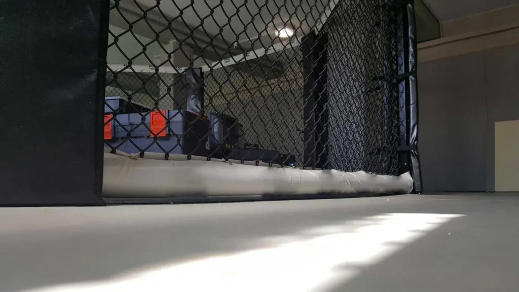 MMA cage frame padding with heavy-duty PVC-covered bottom pad and vertical post pad attached to a chain-link fence.