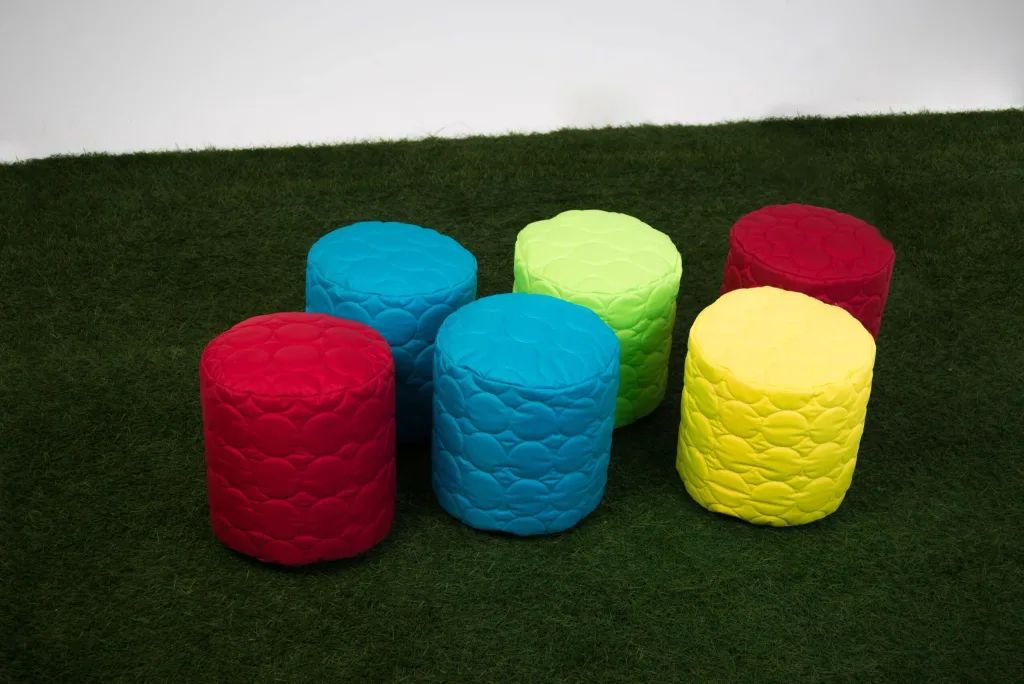 Six small round padded child bean seats, two blue, two red, one yellow and one lime green, arranged on dark green artificial grass against a white wall.