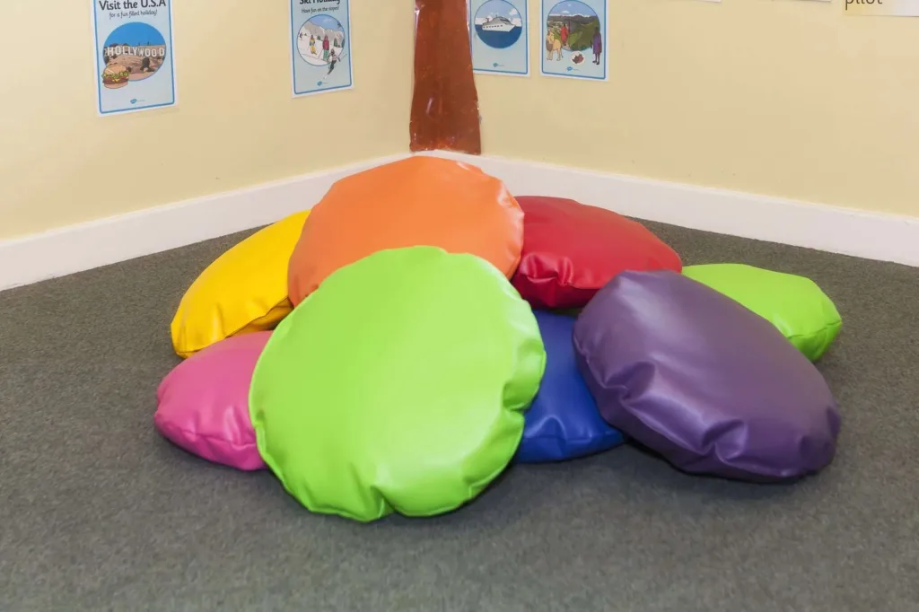 Set of ten round faux leather child scatter cushions in assorted bright colours on a carpeted floor, with a wall poster reading Visit the U.S.A.
