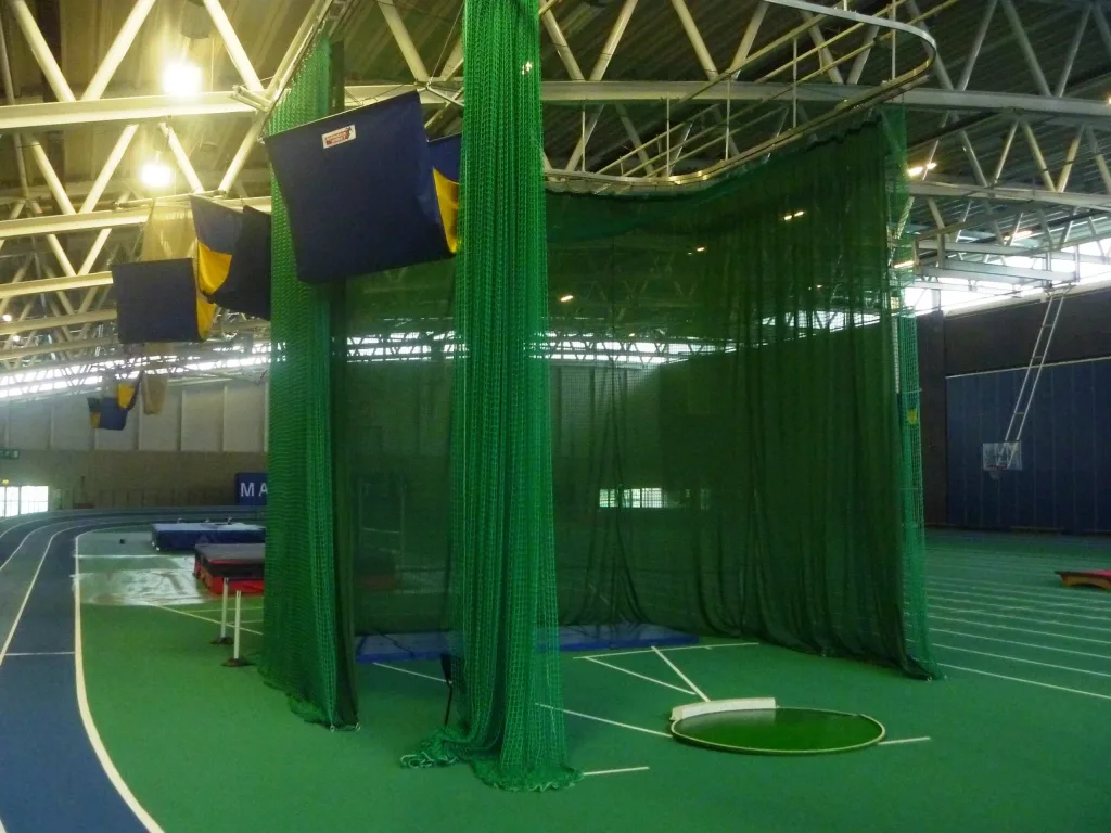 Indoor throws cage with green nets surrounding a throwing circle on a green infield beside a blue indoor running track.