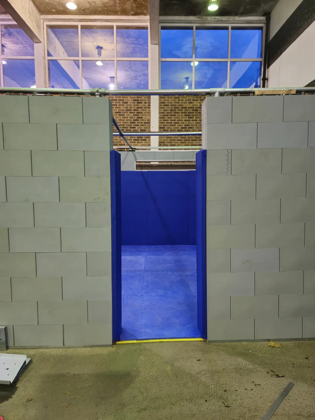 Modular gray block wall with a central doorway showing a blue padded interior and floor inside a high-ceiling warehouse.